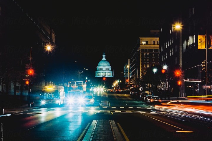 Night city street with car lights and illuminated capitol building in distance.