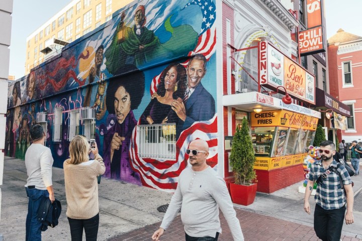 Street mural with diverse figures on wall near Ben's Chili Bowl, people taking photos.