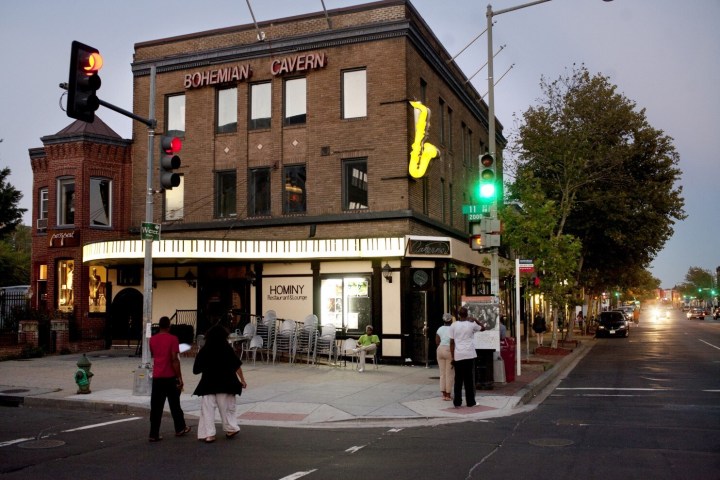 Street corner with a brick building labeled 'Bohemian Cavern' and a neon saxophone sign at dusk.