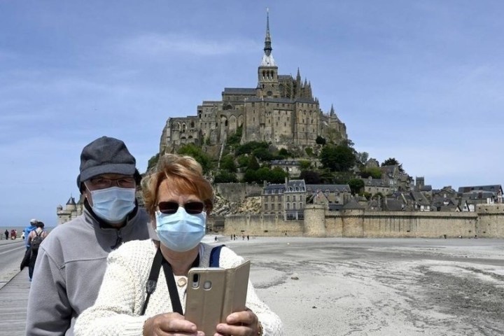 Two people wearing masks taking a selfie in front of Mont Saint-Michel on a cloudy day.