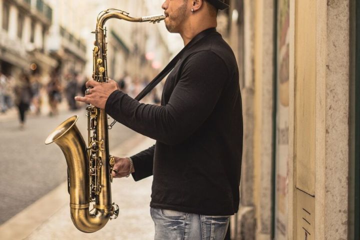 Man playing saxophone on a city street, wearing a hat and casual clothing.