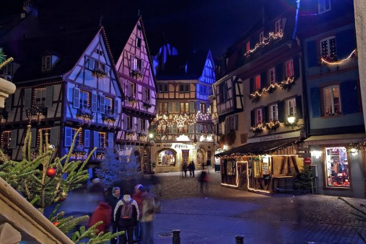Decorated half-timbered buildings with Christmas lights at night, people walking in a festive plaza.