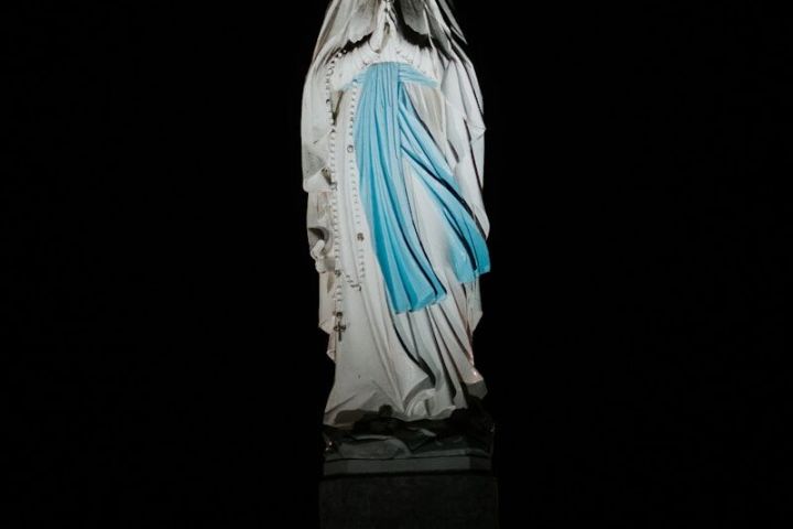 Illuminated statue of crowned figure in white robe with blue sash against night sky.