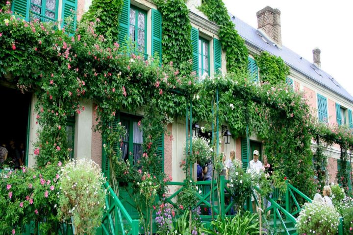 Large house with green shutters and ivy, surrounded by lush garden and flowers, people on porch.