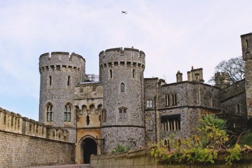 Gray stone castle with two round towers and airplane overhead.