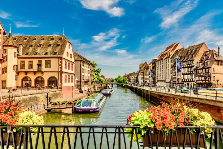 Scenic canal view with historic buildings, two glass-roofed boats, and vibrant flowers under a blue sky.