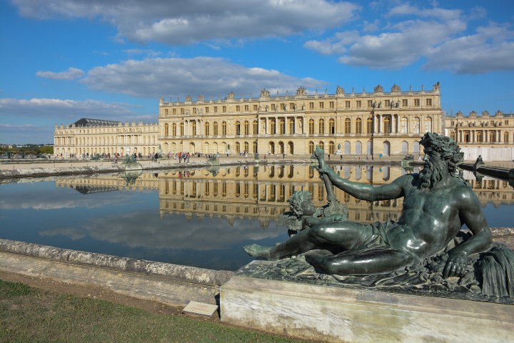 Statue in front of palace with reflection in pond under blue sky with scattered clouds.