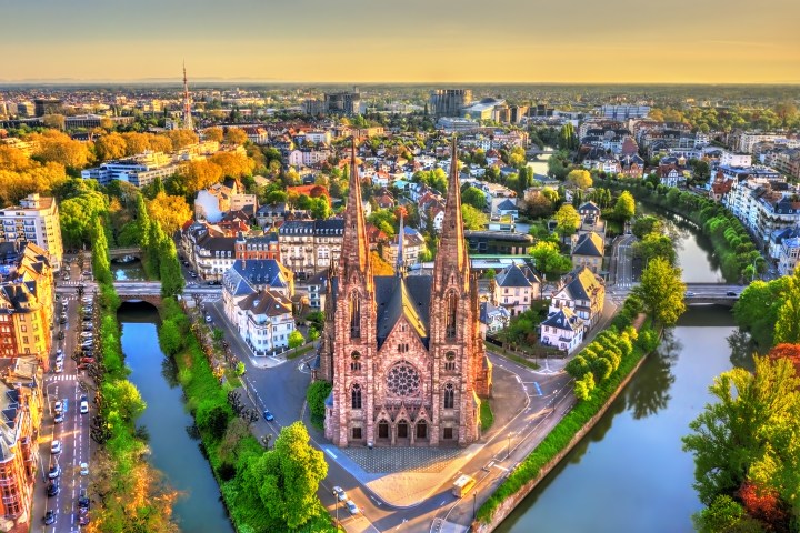 Aerial view of a historic cathedral with twin spires surrounded by a cityscape and river.