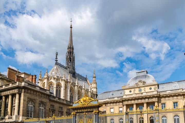 Gothic chapel with tall spire next to an ornate building under a partly cloudy blue sky.