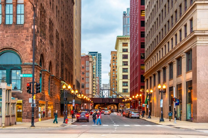 Urban street with people, cars, tall buildings, and overpass under cloudy sky.