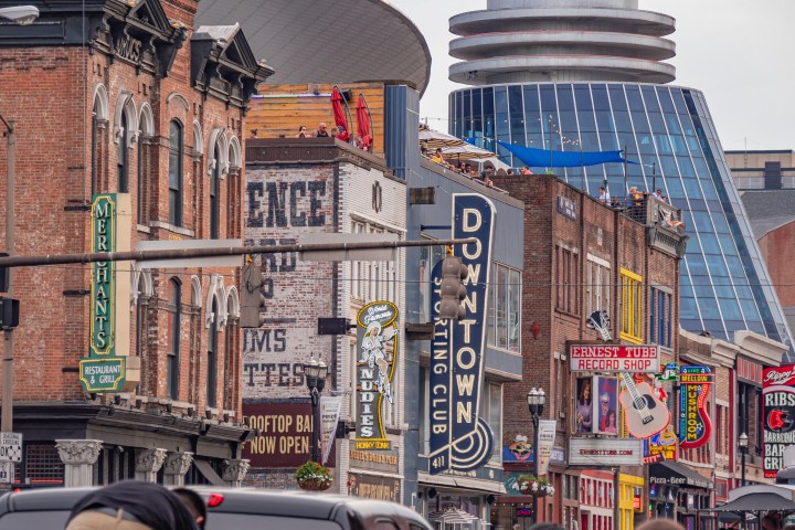 City street with brick buildings, neon signs, and a modern glass structure in the background.