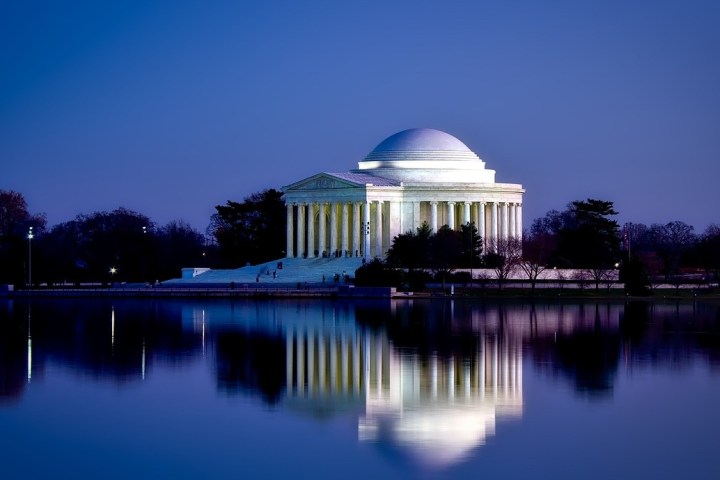 Illuminated building with dome and columns reflected in water at dusk.