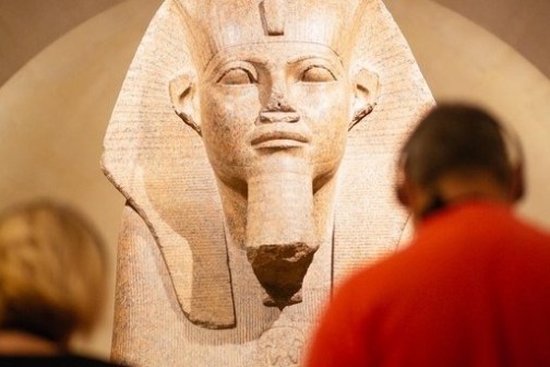 Two people in a museum viewing a large ancient Egyptian statue in a dimly lit room.