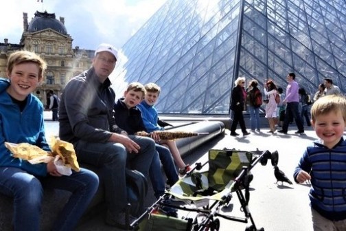 A family eating by the Louvre Pyramid under sunlight, with people walking in the background.
