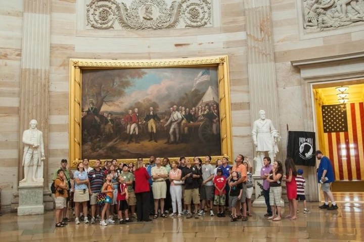 Tourists gather in a historic building with large paintings and statues, near a U.S. flag and POW/MIA flag.