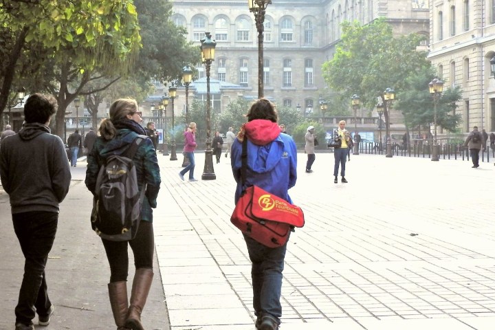 People walking on a tree-lined city street with historic buildings.