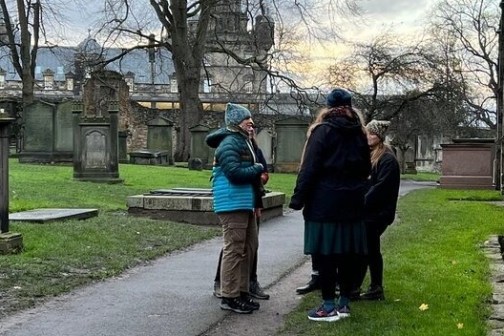 Four people in coats stand talking in a cemetery path surrounded by gravestones and trees.