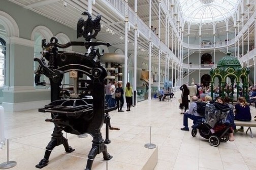 Museum interior with a large ornate metal machine and visitors sitting in a spacious hall.