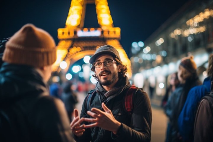 Man in glasses and cap speaks to group near illuminated tower at night.