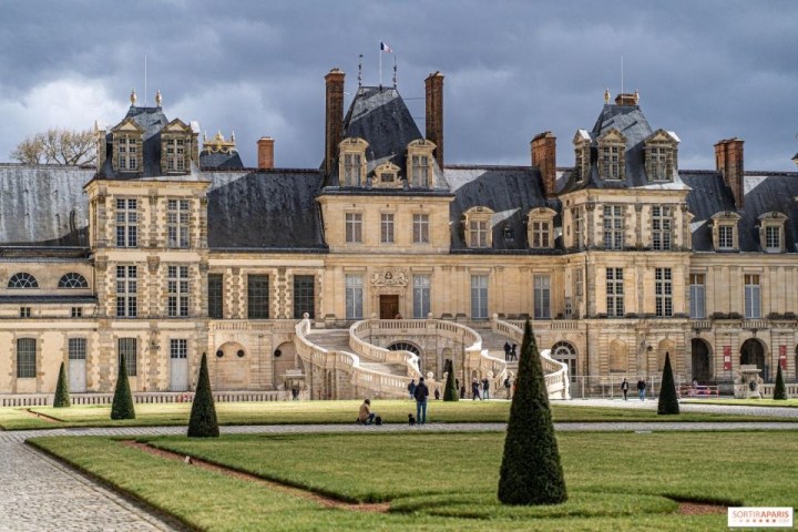 Historic chateau with ornate facade, staircase, and manicured lawn under a dramatic cloudy sky.