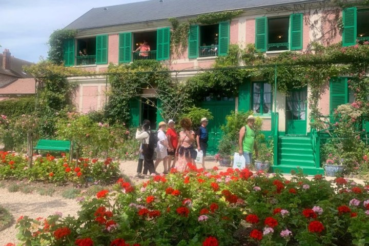 People walking by a house with green shutters, surrounded by vibrant garden flowers and climbing plants.
