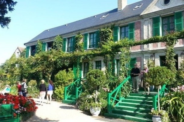 People near a vine-covered house with green shutters and stairs in a garden.