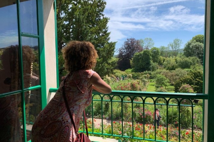 Person with curly hair admiring a lush garden from a veranda on a sunny day.
