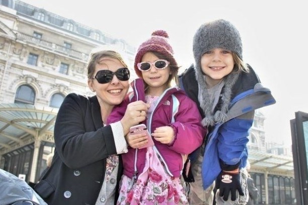 A smiling woman and two children in warm clothes pose outside a historic building.