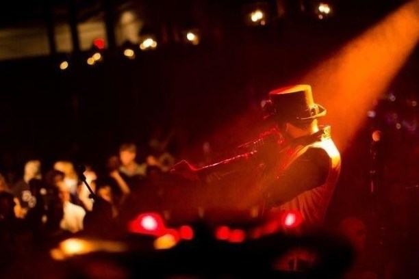 Performer in hat illuminated on stage with spotlight, crowd visible in dim background.