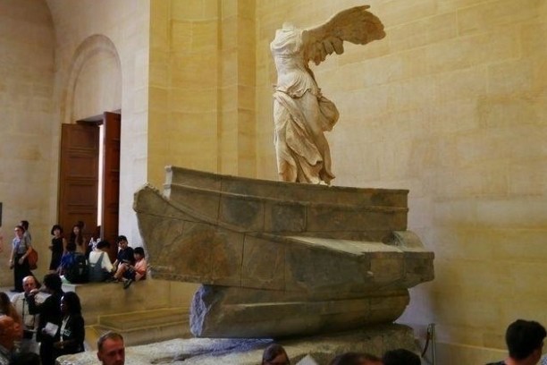 Winged statue on stone pedestal in museum surrounded by visitors.