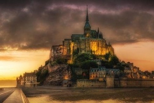 Illuminated Mont Saint-Michel under a dramatic cloudy sunset sky.