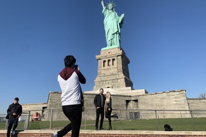 People taking photos near the Statue of Liberty under a clear blue sky.