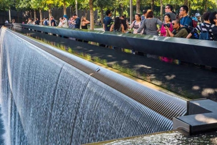 People observe a large, cascading waterfall at a memorial site with trees in the background.