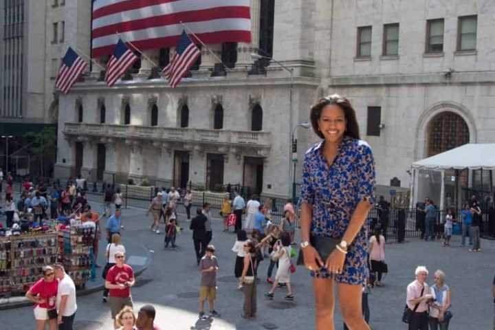 Smiling woman in blue dress stands near NYSE with large American flag in background.