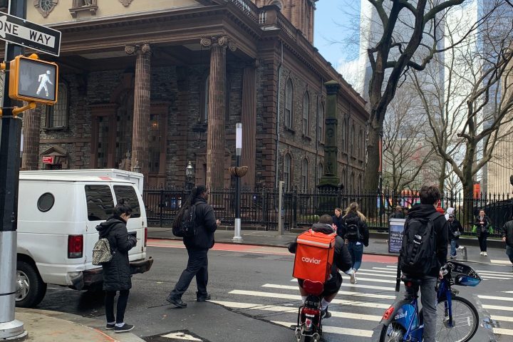 Urban scene with pedestrians and cyclists near historic building and leafless trees.
