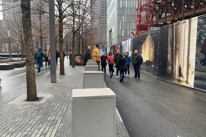 People walking on a city sidewalk beside a construction site with cranes and buildings.