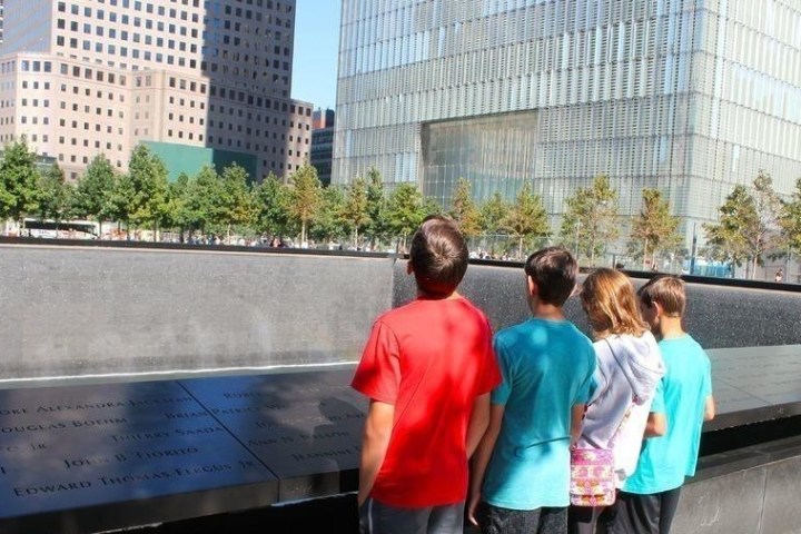 Four children facing a memorial with engraved names, surrounded by urban buildings and trees.