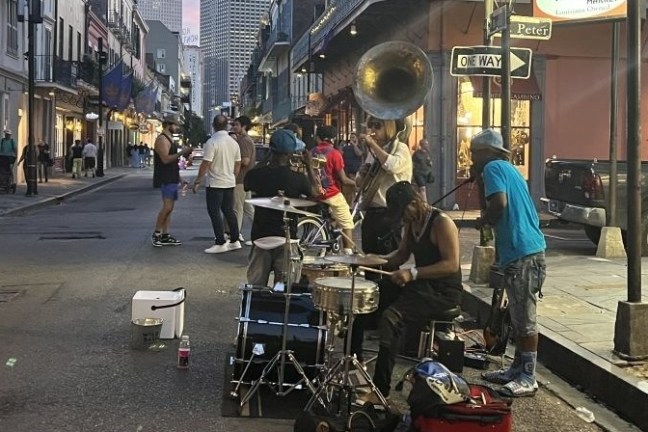 Street musicians play with drums and brass instruments, attracting a small crowd in an urban setting.