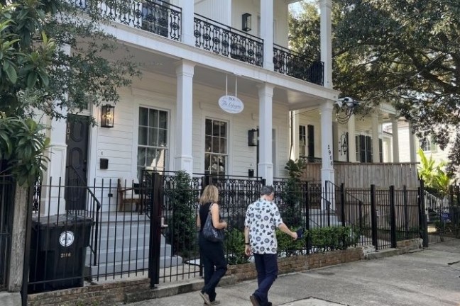 Two people walk past a white two-story house with a porch and trees.
