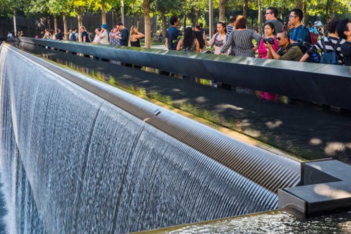 Memorial waterfall with people observing from a railing under tree shade.