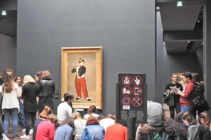 People gathered in front of a painting in a museum gallery with a sign showing prohibited actions.