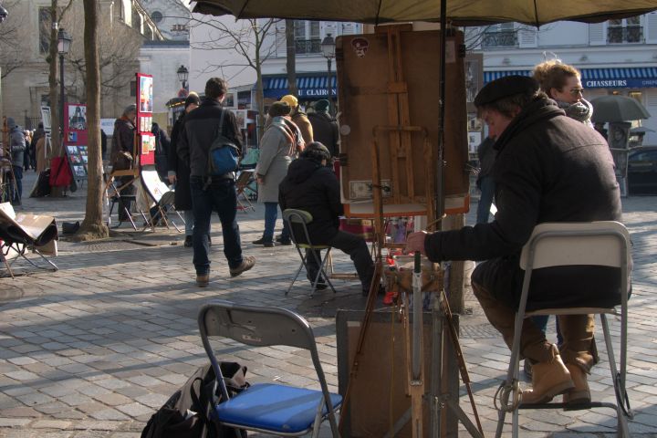 Outdoor artists paint and display art on an urban cobblestone street, people walking by.