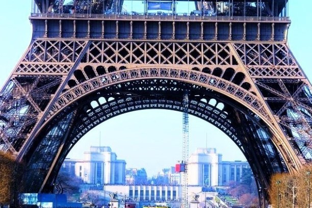 Base of the Eiffel Tower with a clear blue sky and distant buildings visible through the arch.