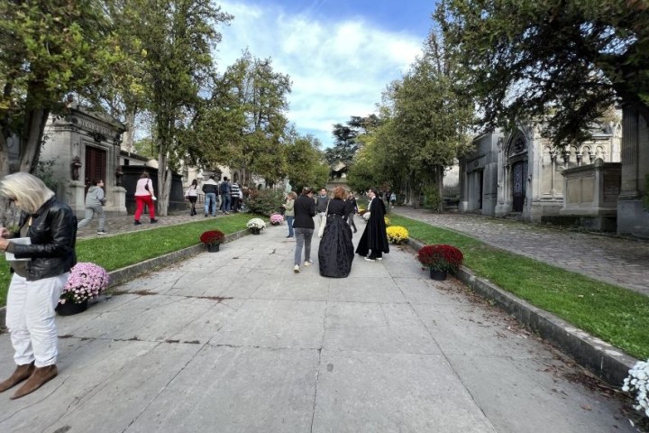 People walking in a cemetery with trees and flowers on a sunny day.