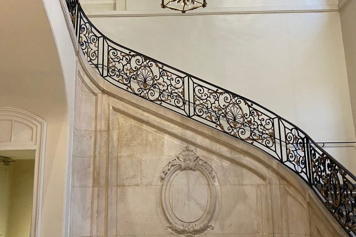 Elegant staircase with ornate railing, chandelier, and a row of white chairs on a checkered floor.