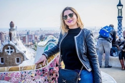 Woman in sunglasses and leather jacket smiling at Park Güell with cityscape in the background.