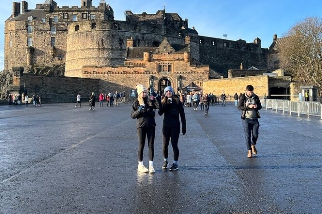 People walking in front of an old stone castle under a clear blue sky.
