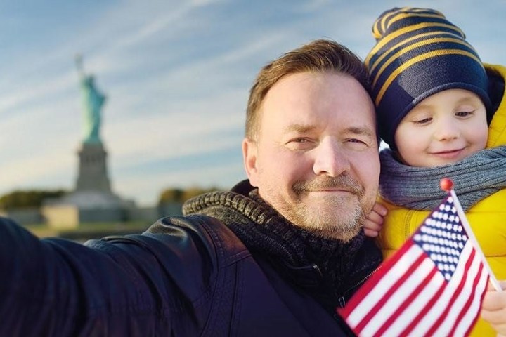 Man and child selfie with American flag, Statue of Liberty blurred in background.