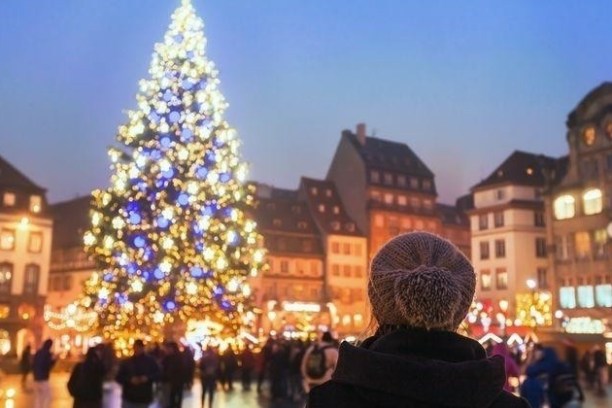 Person in winter wear facing a large, decorated Christmas tree in a city square.