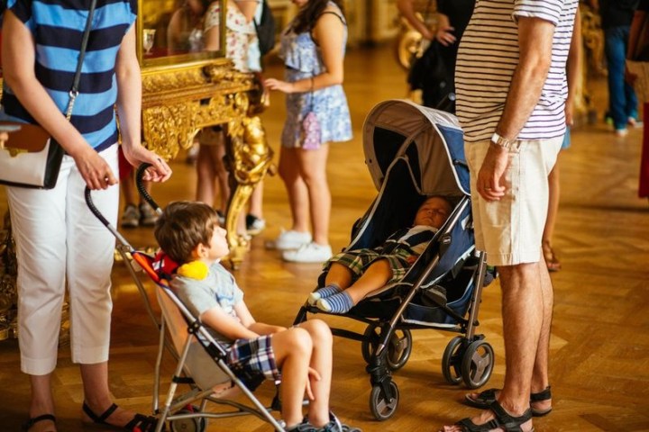 Two children in strollers indoors, surrounded by adults and ornate furniture.